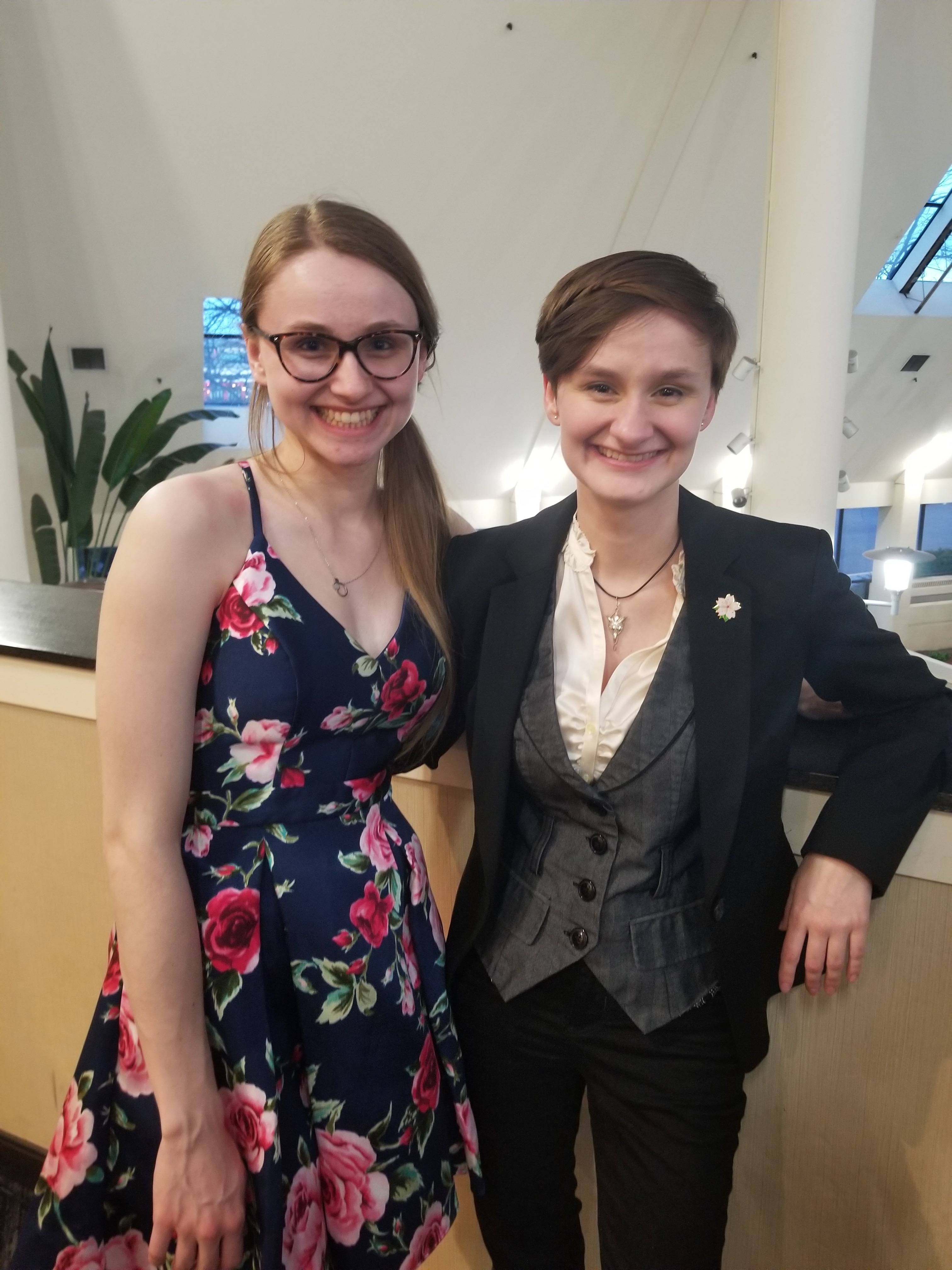 Two young women dressed up as wedding guests stand arm in arm smiling at the camera