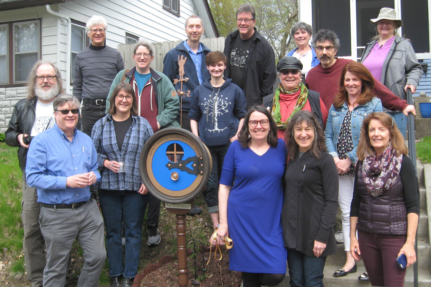 A group of people stand in front of a house surrounding a new hobbit-hole shaped Little Free Library with a blue door.