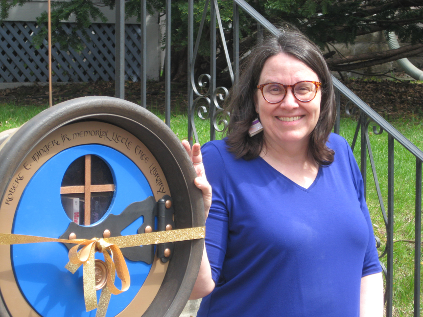 A woman stands, smiling, besides a hobbit-door shaped Little Free Library. The library is enclosed by a gold ribbon