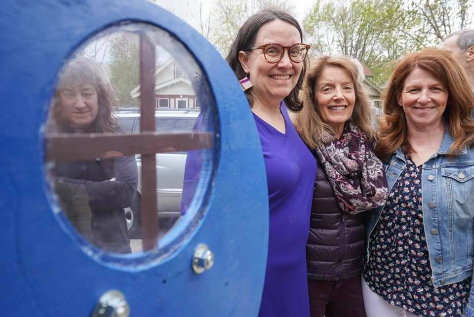Picture taken from inside Little Free Library shows three women (Peg and her sisters Betsy and Cindy) standing beside it outside. Another friend can be glimpsed through the Little Free Library window