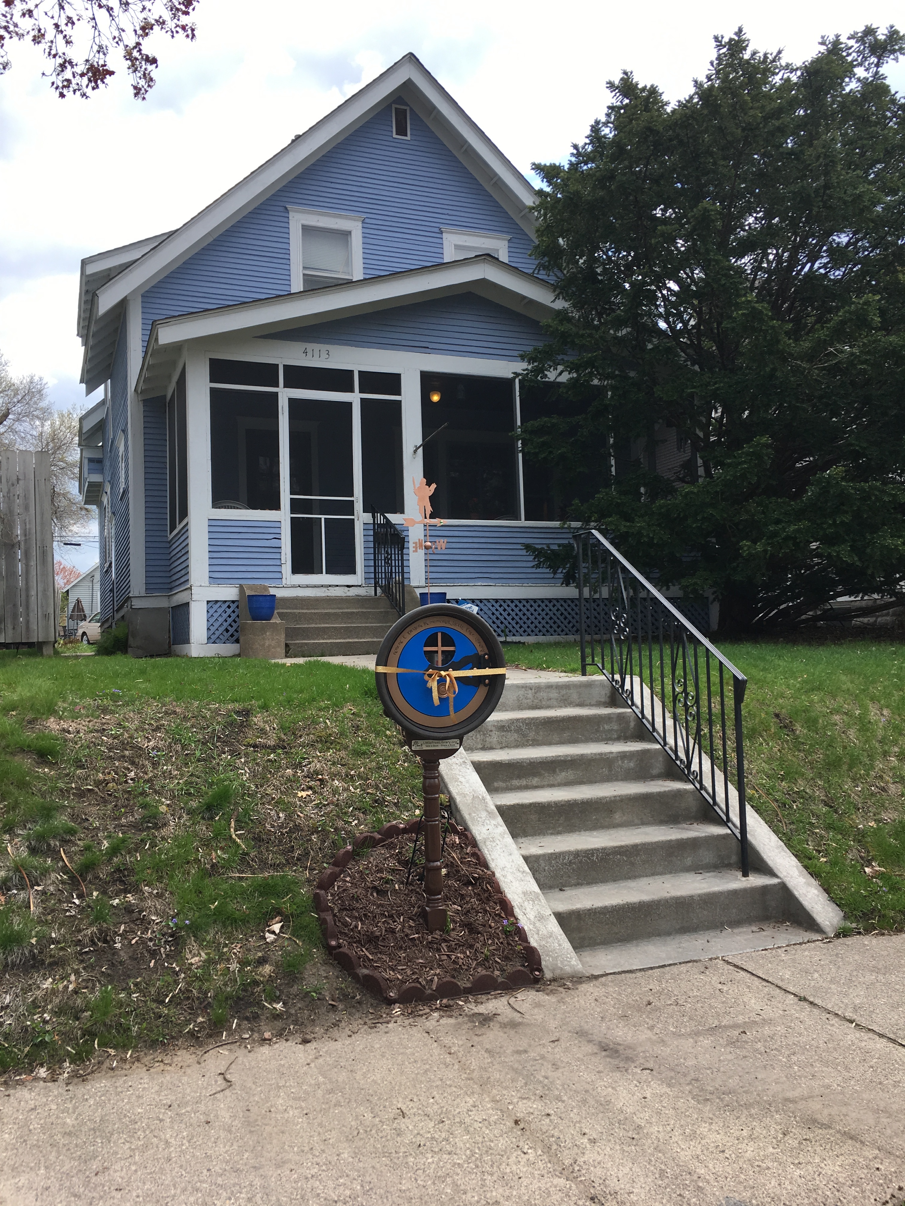 Blue house with white trim up on a hill. Besides the stops leading up to the house is a round hobbit-door shaped Little Free Library with a blue door.
