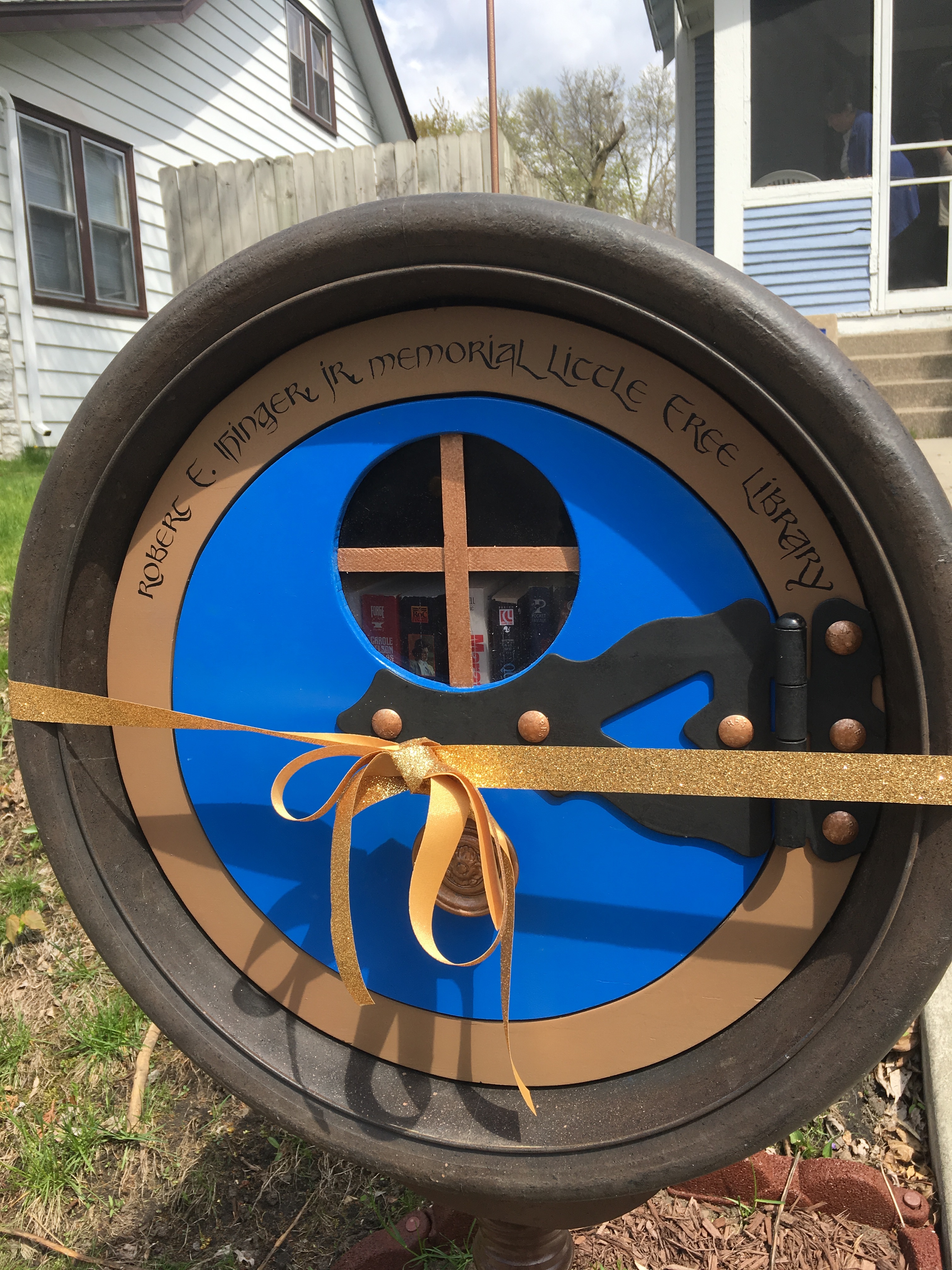 Closeup of a round hobbit-door shaped Little Free Library. Above the door are the words "Robert F. Ihinger, Jr. Memorial Little Free Library." A gold ribbon is wrapped around the library tied with a bow in front.