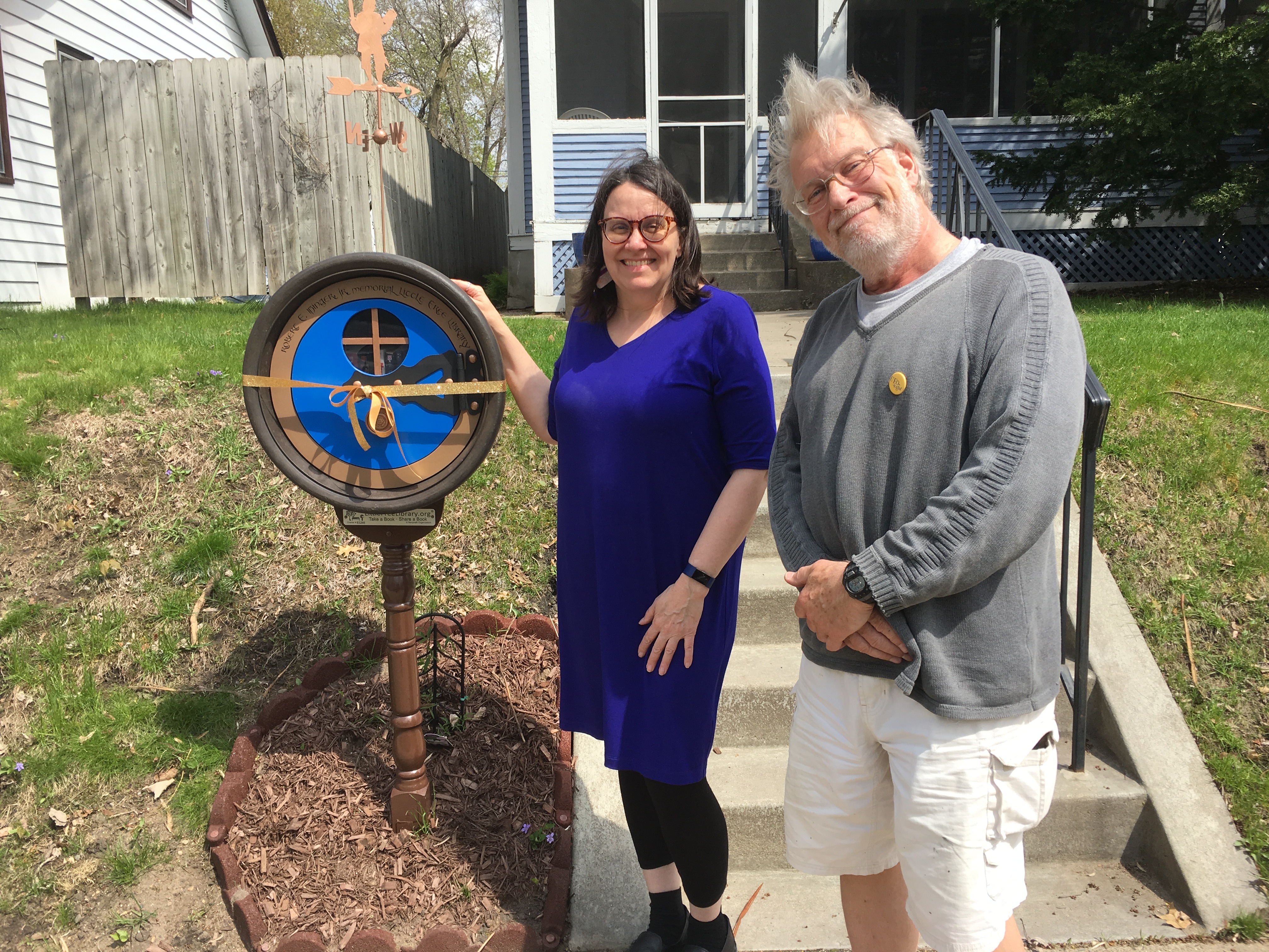 A woman (Peg) and a man (Terry Faust, the designer) stand in front of a Little Free Library in front of a house.
